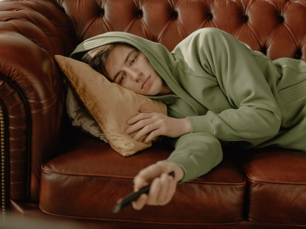 A bored guy in a hoodie watches tv lying on a couch. Photo by cottonbro from Pexels https://www.pexels.com/photo/woman-in-gray-hoodie-lying-on-brown-leather-couch-4114908/